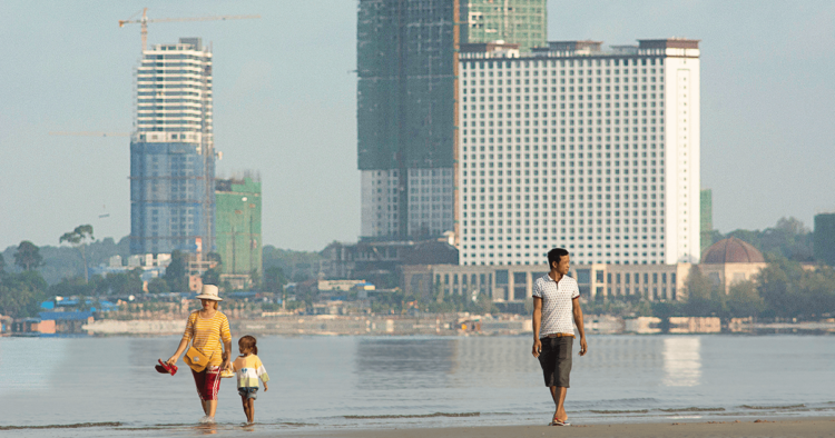 A brightly dressed woman and child, and further off, a man, walked on the beach in front of a backdrop of shiny new construction.