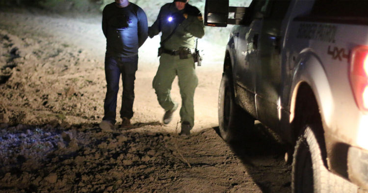 A Border Patrol agent walks next to a man in handcuffs by the side of a patrol truck