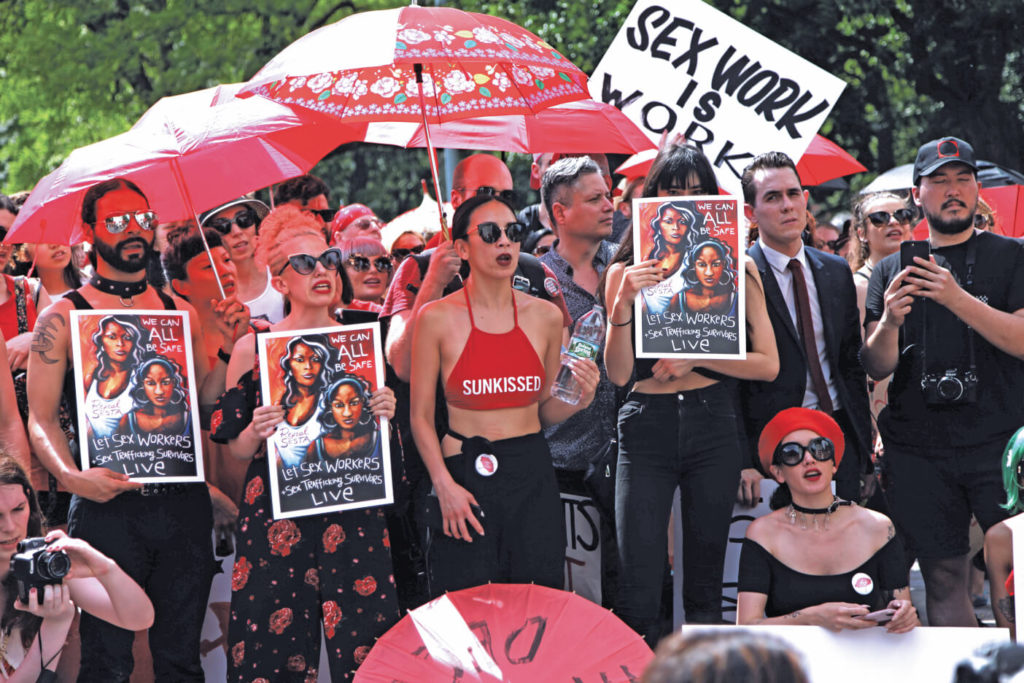 A group of people dressed in red and black hold protest signs beneath umbrellas. One reads "Sex work is work."