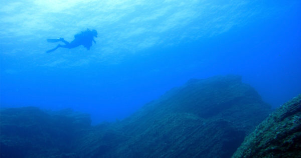A scuba diver descends to the jagged edge of a rock beneath the deep blue sea