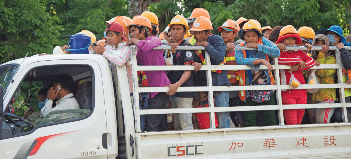 A white truck with an open back filled with yellow-hatted construction workers of all ages and genders.