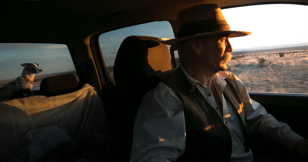 A man and his dog sit in the cab of a druck, with the sun filtering in over ranchland.