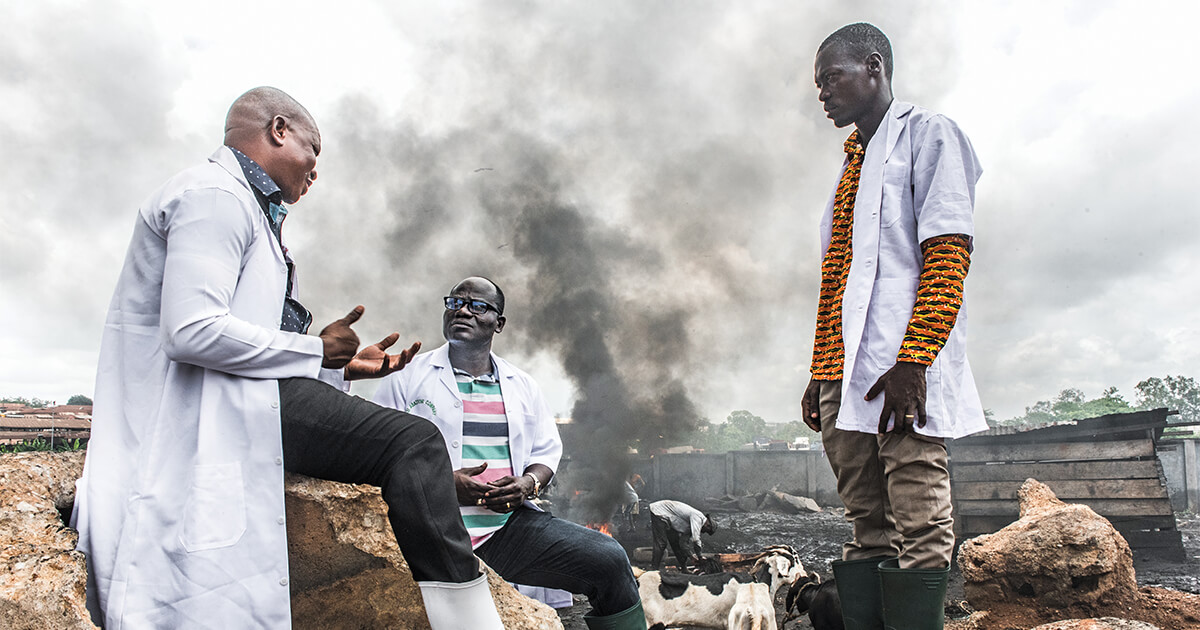 Three men talking in a meat processing facility in Ghana as tire fires burn in the background