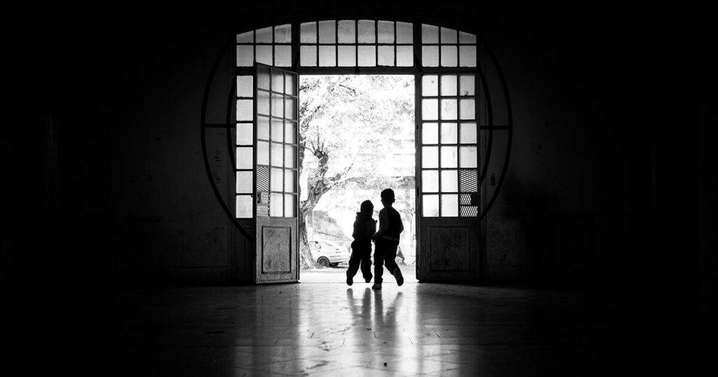 A black-and-white photograph of the silhouettes of two children run towards an open, sunlit door