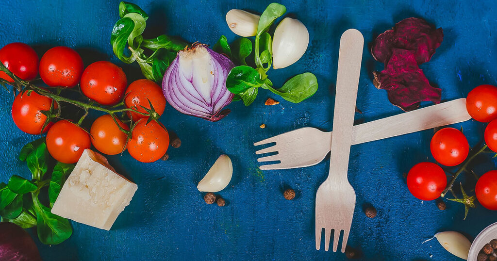 Vegetables, mainly cherry tomatoes, sliced red onion, garlic, and basil, spread out on a blue background. Two wooden forks are crossed among the vegetables.